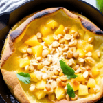 Photo of a dutch baby pancake topped with pineapple, toasted coconut, macadamia nuts and a little bit of mango in a cast iron skillet. S A lime is next to the skillet